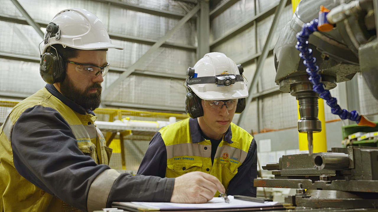Two workers wearing hard hats, safety glasses, and yellow hi-vis stand by a drilling machine. One of the workers appears to be instructing the other, who is looking at a document intently.