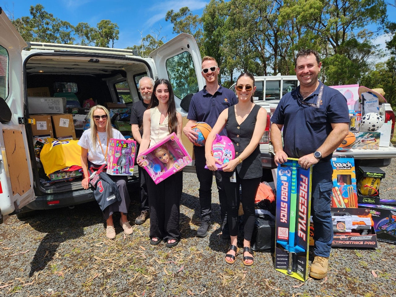A group of six people (one sitting, five standing) are seen outdoors in front of an open white van filled with boxes and bags. They are holding various toys and gifts, including a basketball, pogo sticks, and boxed dolls. Additional toys and items are visible on the ground and in the van. The background shows trees and a clear blue sky.