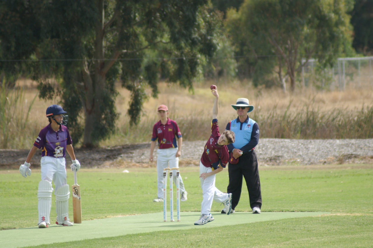 A youth performs an overhead bowl on a cricket field while a young batter watches from the left.