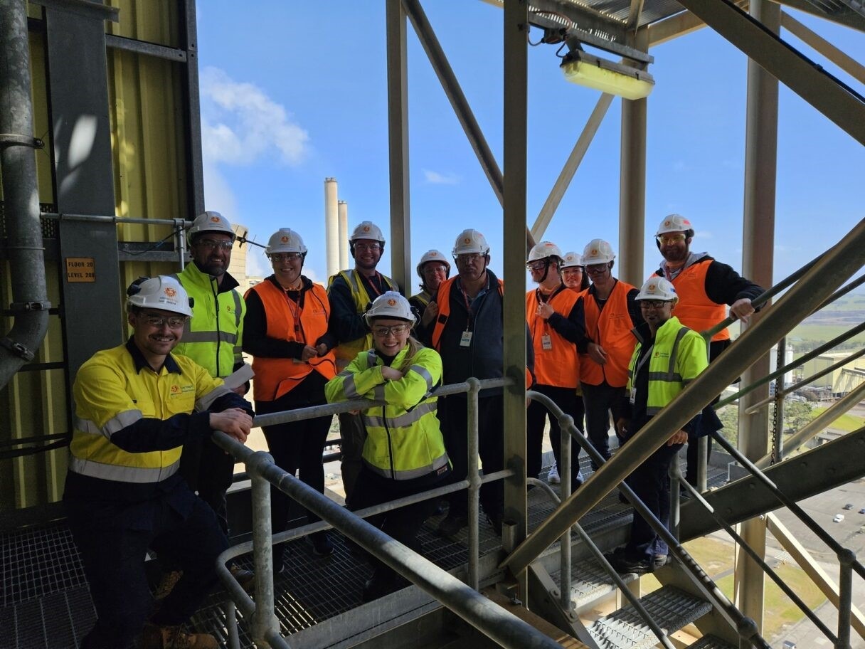 A picture of Interchange Gippsland participants plus mentors dressed in hi-vis, hard hats and safety glasses stand on a staircase at Loy Yang B Power Station with a view of the Gippsland behind them.