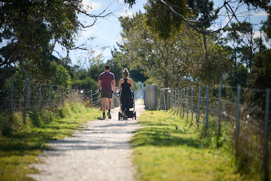The backs of two people walking on the Gippsland Plains Rail Trail. The woman is pushing a pram, the man is walking a dog.