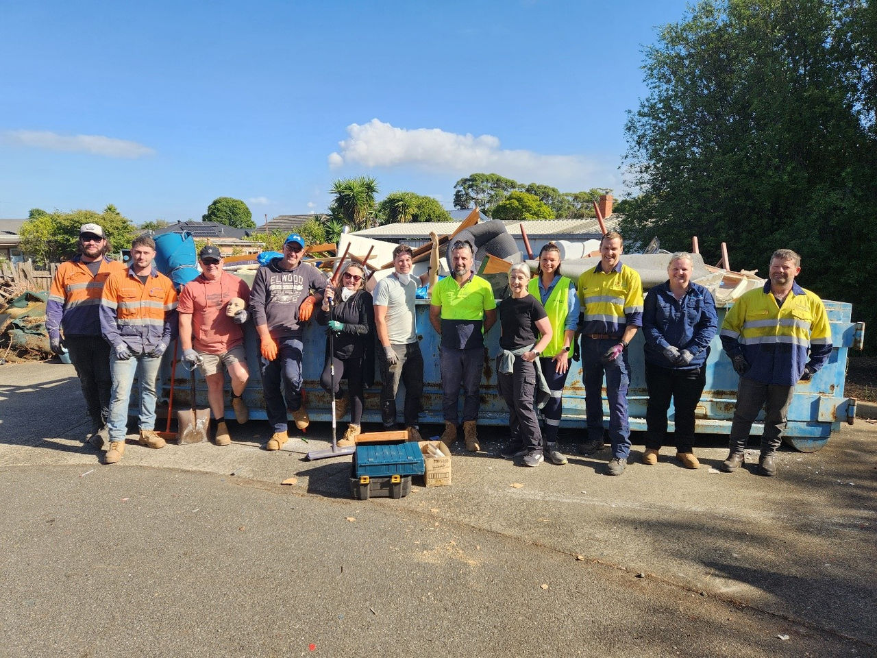 Group of volunteers standing outdoors beside a large skip filled with collected items, smiling after a community clean‑up effort