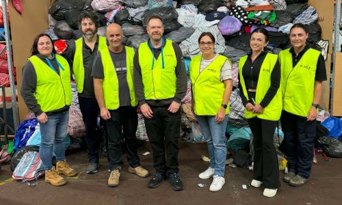 A group of workers in hi-vis clothing standing in front of a large backlog of donations for Lifeline Gippsland.