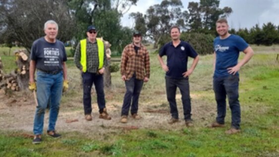 Five people stand on a grassy rural clearing beside a stack of cut logs, wearing casual work clothing; trees and open paddock extend into the background under an overcast sky.