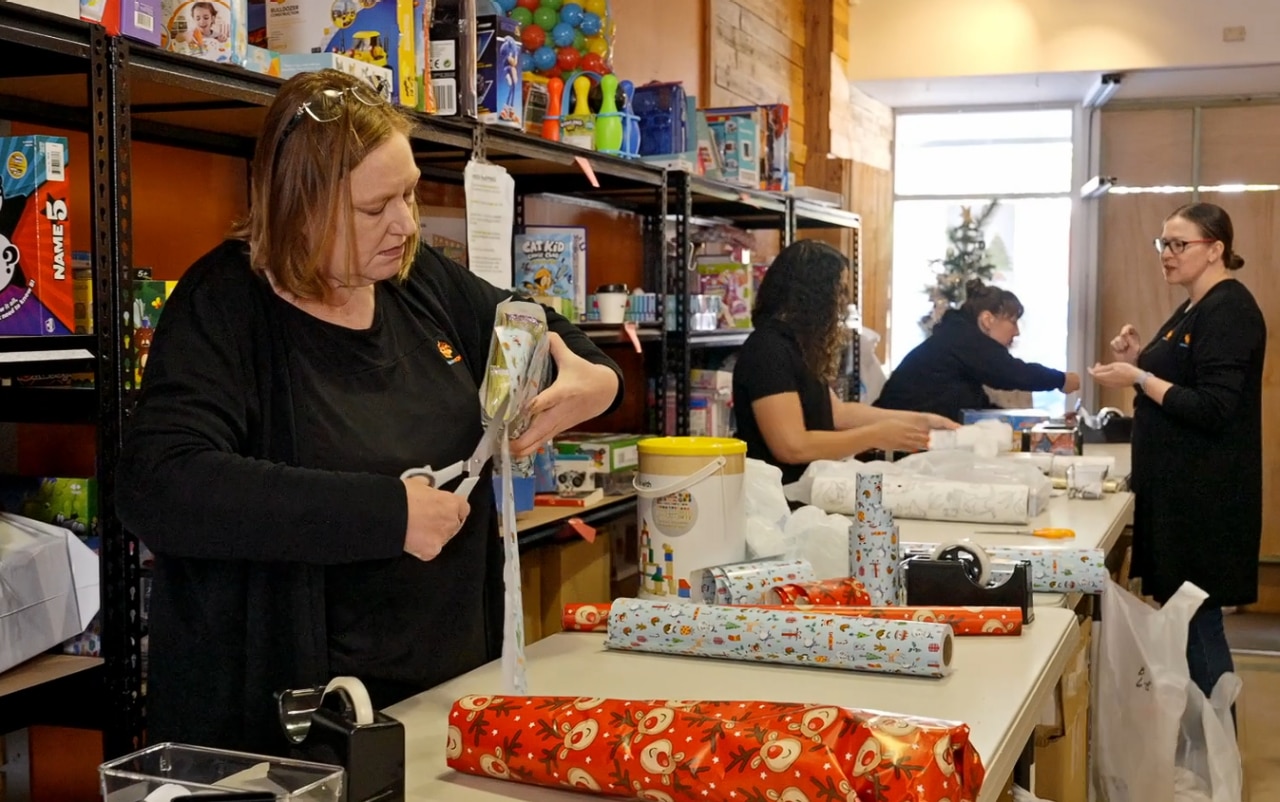 A woman in a black shirt uses a pair of scissors while wrapping presents with several colleagues.